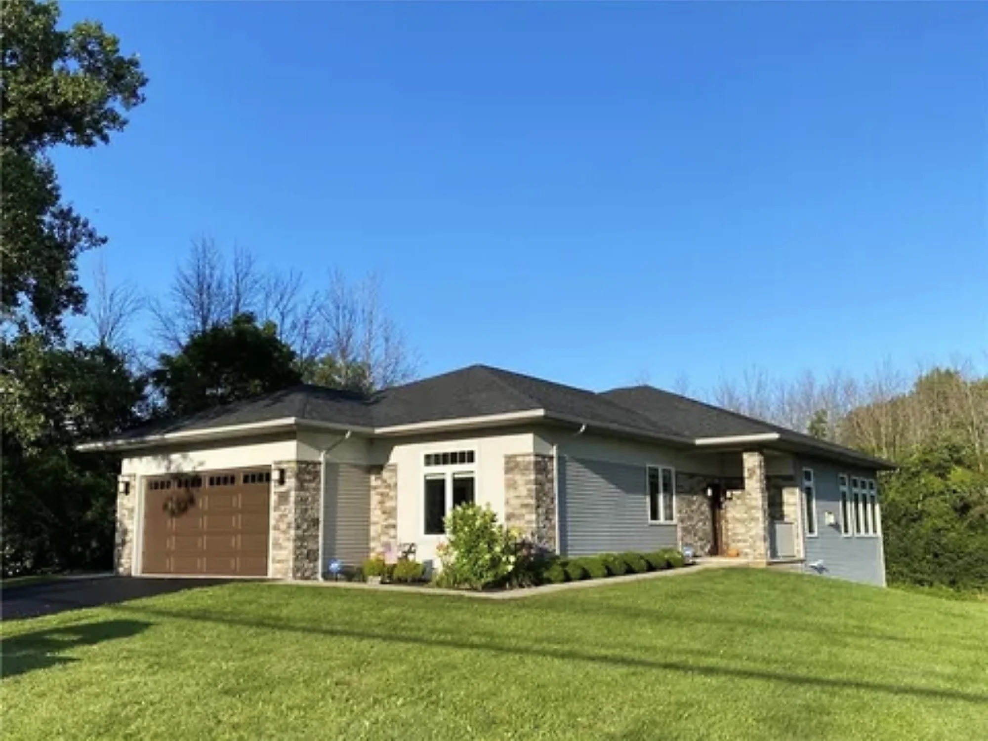 The front of a single-story home with a dark shingled roof, gray siding, stone columns, and a two-car garage.